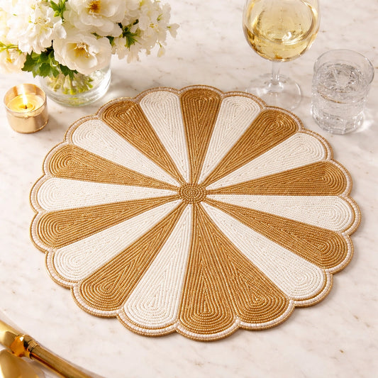 A round woven placemat with a scalloped edge, featuring a radial fan pattern in tan and off-white. It is styled on a white marble tabletop with a glass of white wine, a glass of water, a candle, and white flowers.