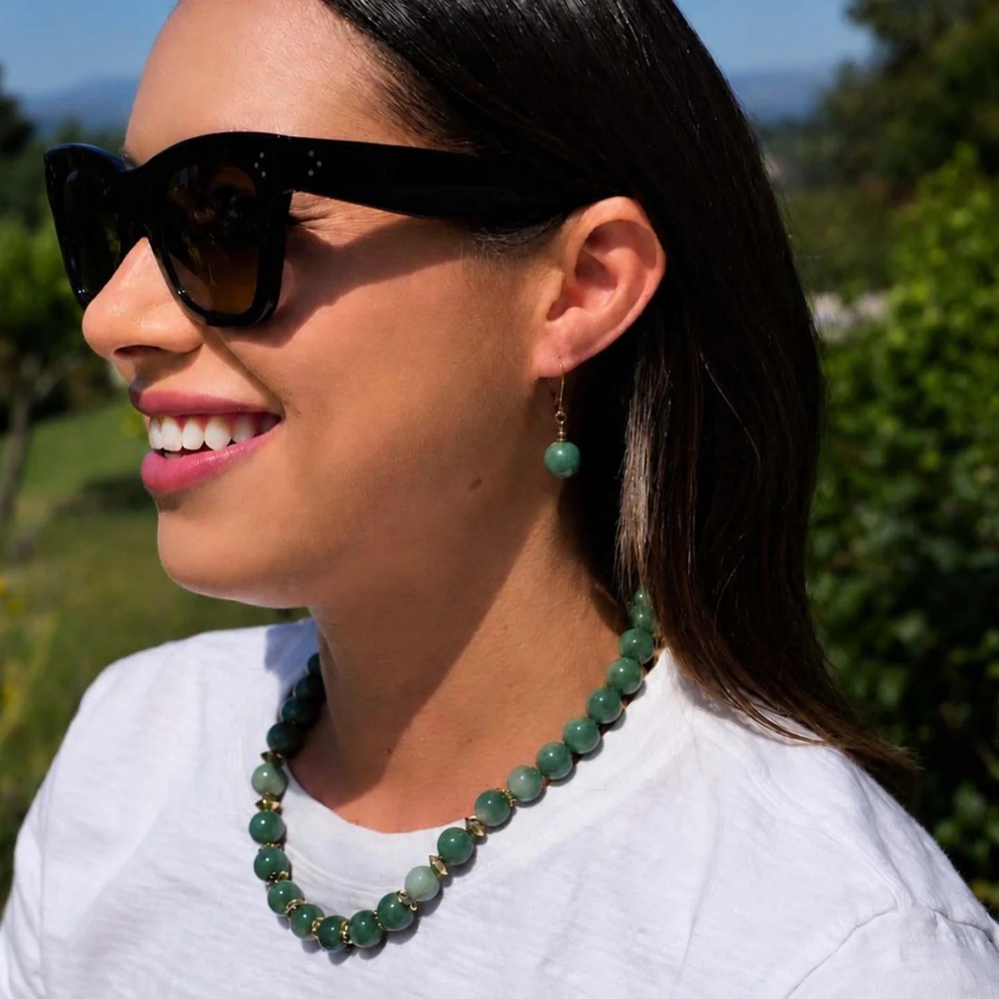 A woman outdoors wearing a green beaded necklace with gold spacer beads, matching green drop earrings, and black sunglasses.