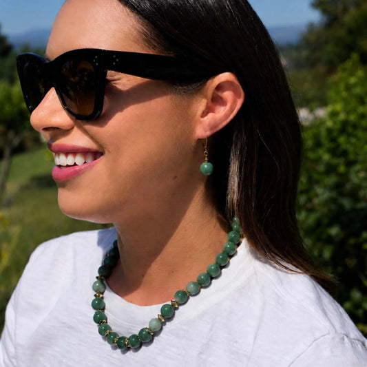 A woman outdoors wearing a green beaded necklace with gold spacer beads, matching green drop earrings, and black sunglasses.