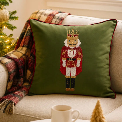 An olive green throw pillow with a red-trimmed border and a sequined nutcracker figure, styled on a beige couch with a plaid blanket and holiday decor.