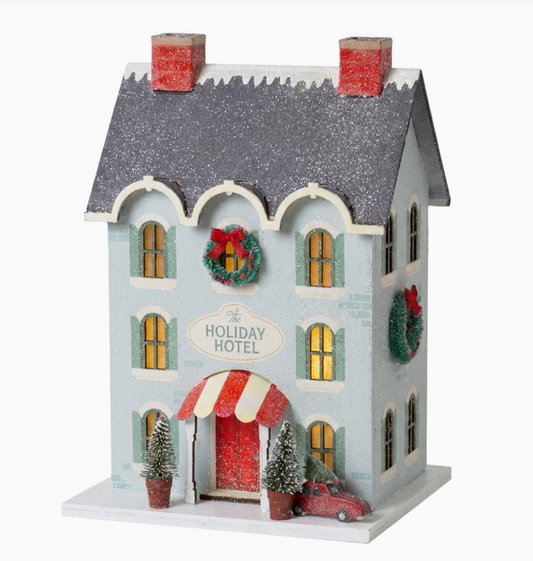 Lighted Holiday Hotel Christmas village decor in blue with glittered gray roof, red door, festive wreaths, miniature trees and holiday car.
