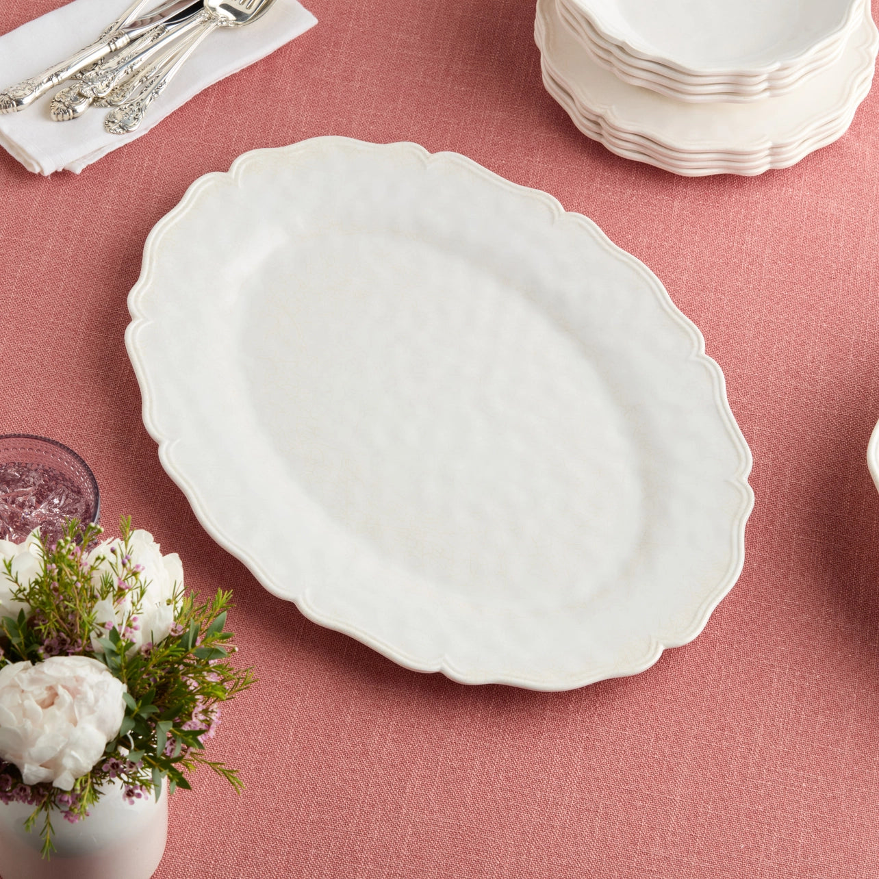 White scalloped-edge plate on a pink tablecloth with floral arrangement and silverware.