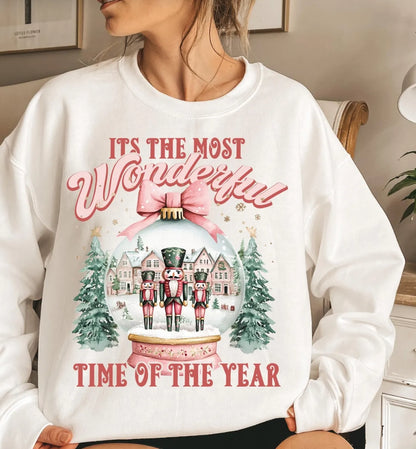 A woman wearing a white crewneck sweatshirt with a festive Christmas graphic that reads 'IT'S THE MOST Wonderful TIME OF THE YEAR' around an illustration of nutcrackers in a snow globe with trees and houses.