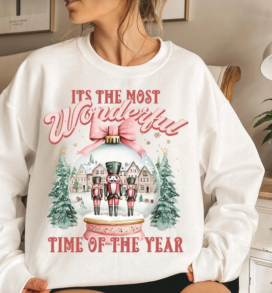 A woman wearing a white crewneck sweatshirt with a festive Christmas graphic that reads 'IT'S THE MOST Wonderful TIME OF THE YEAR' around an illustration of nutcrackers in a snow globe with trees and houses.
