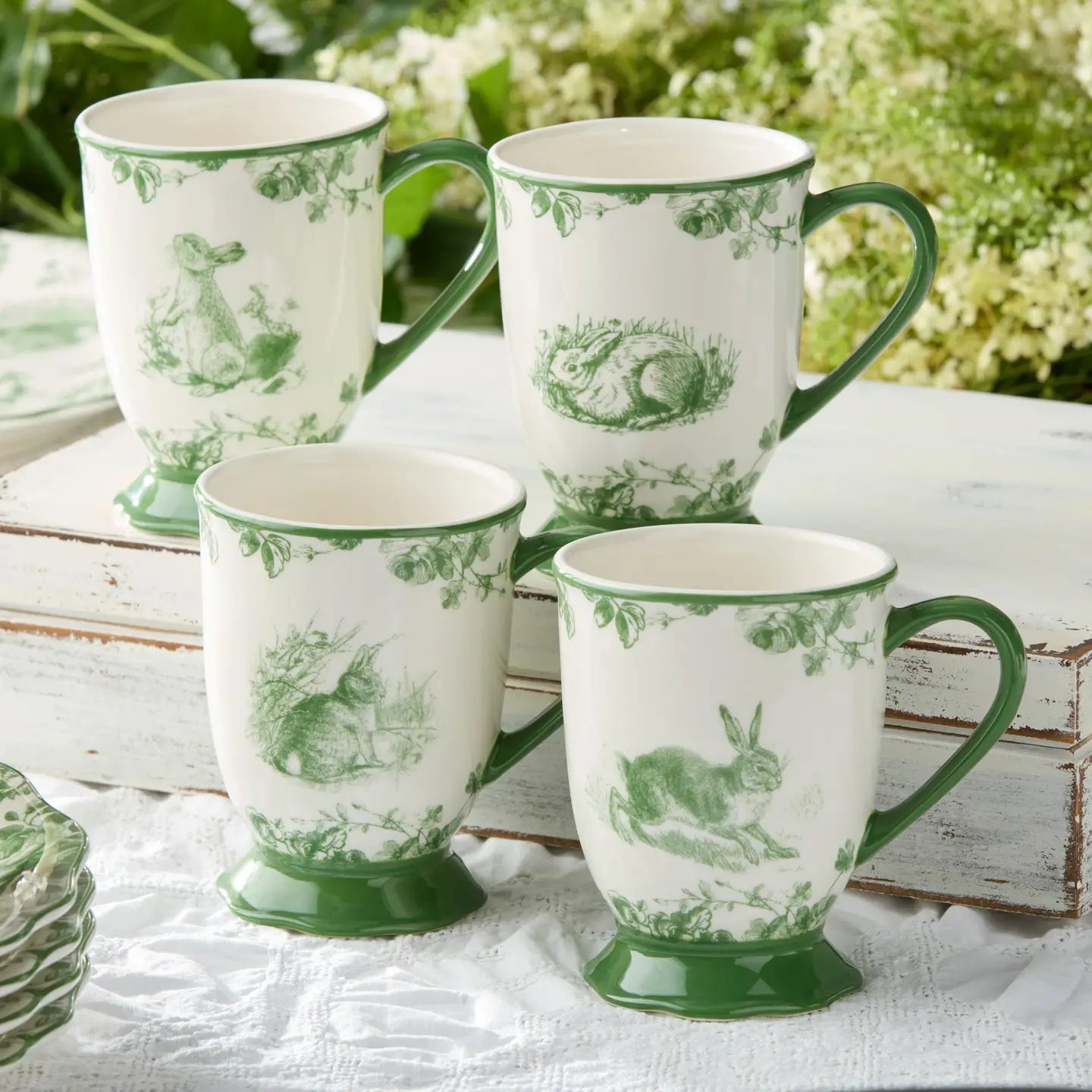 Set of four white mugs with green floral patterns on a white surface with greenery in the background.