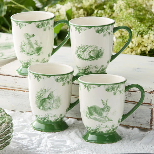 Set of four white mugs with green floral patterns on a white surface with greenery in the background.