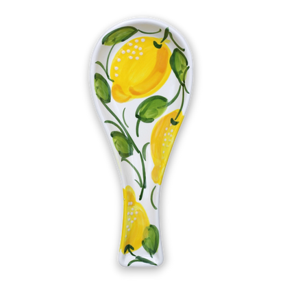 A white ceramic spoon rest decorated with a hand-painted style yellow lemon and green leaf pattern, shown on a clean white background.