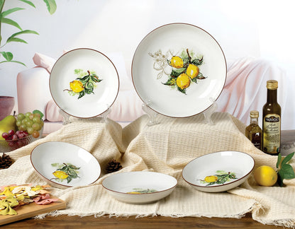 A set of white ceramic plates and bowls decorated with yellow lemon and green leaf illustrations, styled on a beige woven cloth with lemons, grapes, olive oil bottles, and a small cutting board in a bright kitchen setting.