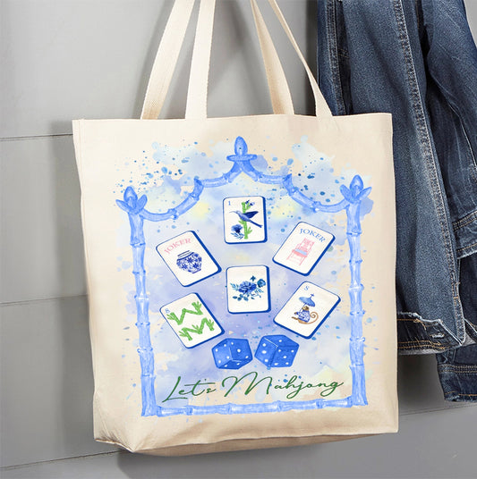 A cream canvas tote bag with a blue watercolor-style frame and illustrated mahjong tiles and dice, hanging against a gray wall next to a pair of blue jeans.