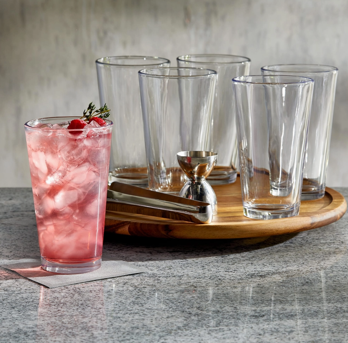Clear glasses on a wooden tray with a pink cocktail and ice cubes on a gray surface.