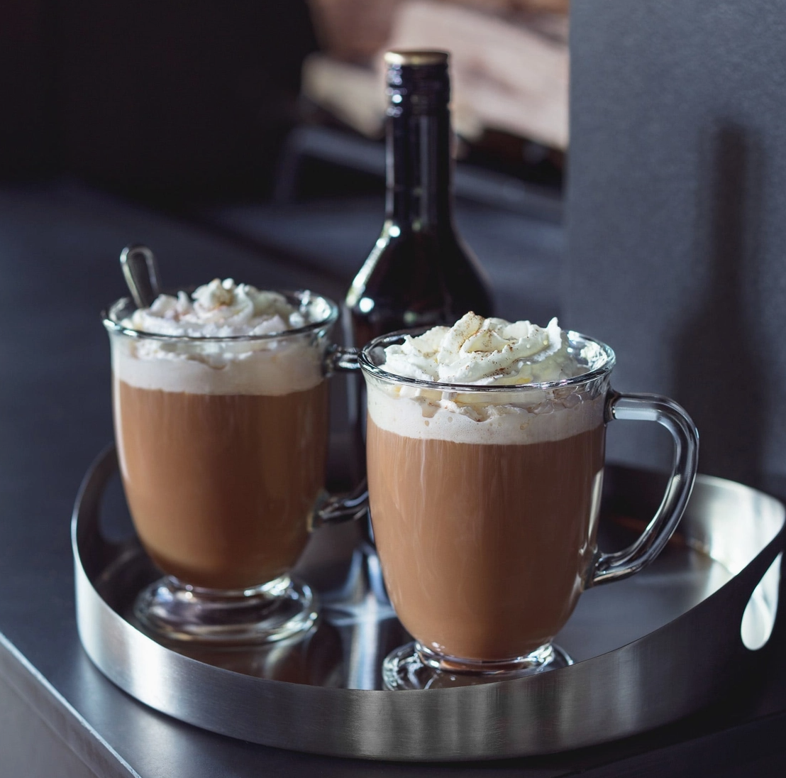 Two glasses of hot chocolate with whipped cream on a metal tray, with a bottle in the background.