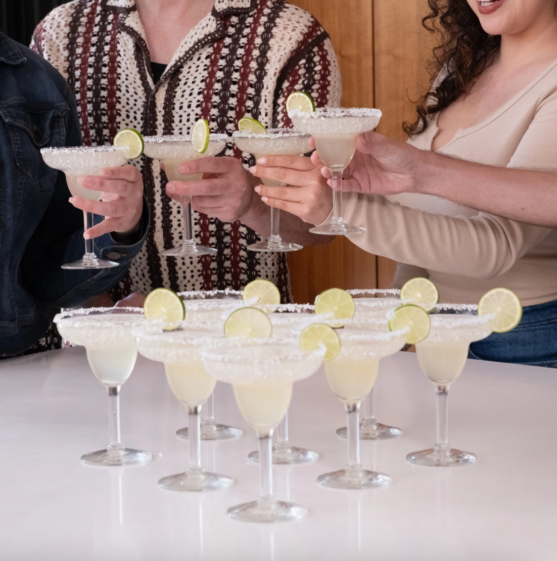 People holding up margarita glasses with lime wedges, with a tray of additional margaritas in the foreground.