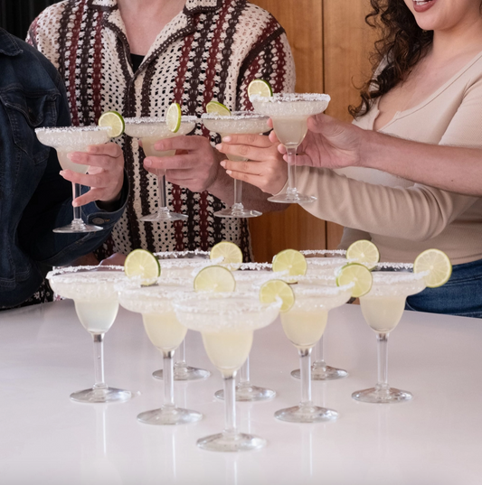 People holding up margarita glasses with lime wedges, with a tray of additional margaritas in the foreground.