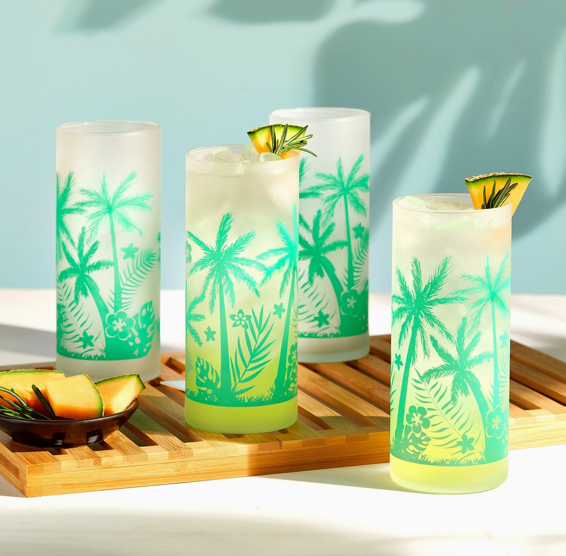 Four glasses with tropical designs on a wooden tray against a light blue background