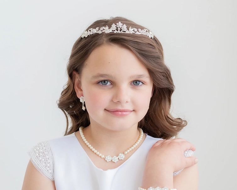 Young girl wearing a white dress with lace sleeves, pearl necklace, bracelet, and communion tiara against a light background.