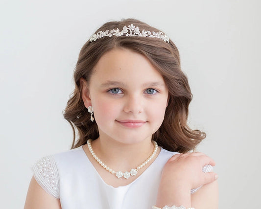 Young girl wearing a white dress with lace sleeves, pearl necklace, bracelet, and communion tiara against a light background.