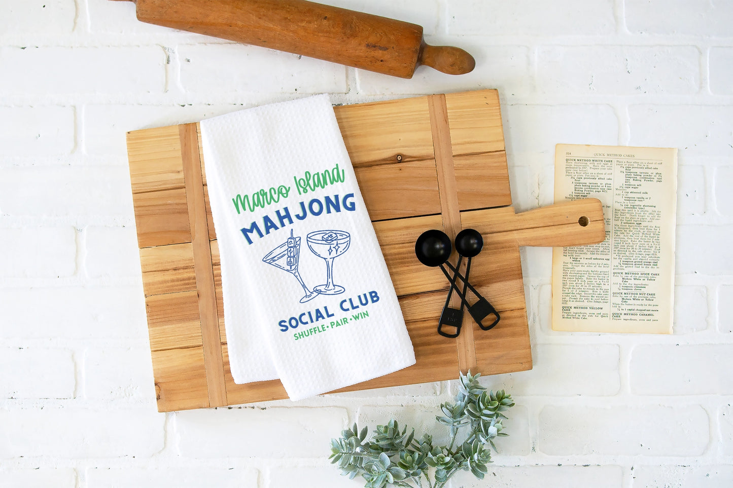 A white kitchen towel printed with 'Majestic Mahjong Social Club' in green and blue, styled on a wooden cutting board with a rolling pin, measuring cups, and a recipe card against a white brick wall.