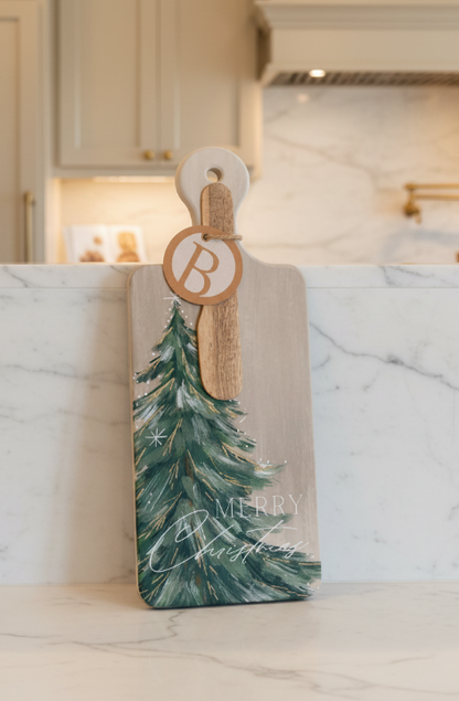 A rectangular wooden serving board with a rounded handle, featuring a painted green Christmas tree and the words “Merry Christmas” on a light wood background. A round wooden tag with a monogram letter “B” is attached with twine.