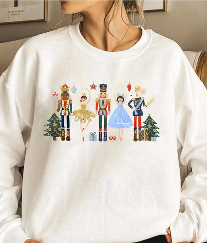 A person wearing a white crewneck sweatshirt with a colorful graphic of nutcracker and ballerina characters standing in front of Christmas trees. NY Gift Boutique
