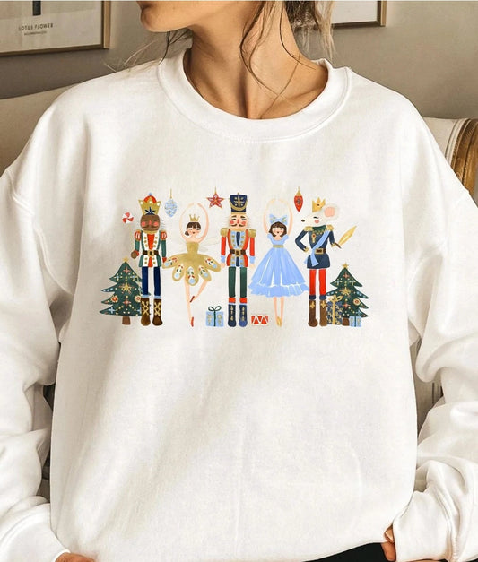 A person wearing a white crewneck sweatshirt with a colorful graphic of nutcracker and ballerina characters standing in front of Christmas trees. NY Gift Boutique