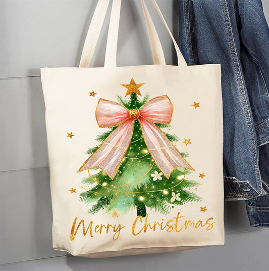 An off-white canvas tote bag with a printed Christmas tree design, hanging next to a blue denim jacket against a gray paneled wall. The bag features a green Christmas tree with a large pink bow, gold stars, and 'Merry Christmas' written in gold script at the bottom.