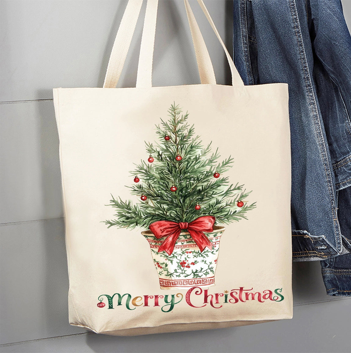 A cream canvas tote bag with a Christmas tree illustration and 'Merry Christmas' text, hanging next to a blue denim jacket against a gray paneled wall.