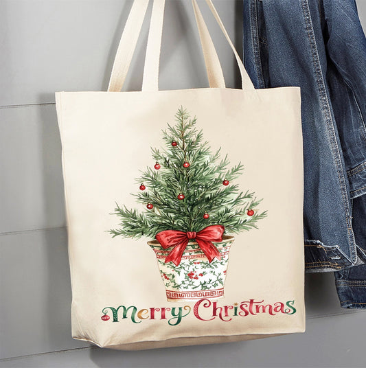 A cream canvas tote bag with a Christmas tree illustration and 'Merry Christmas' text, hanging next to a blue denim jacket against a gray paneled wall.