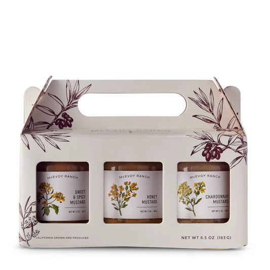 Three mustard jars in a decorative cardboard carrier with floral designs on a white background.