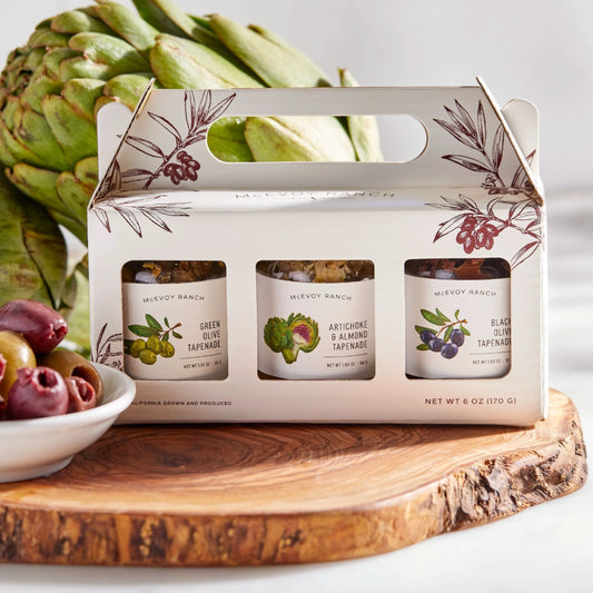 Three tin canisters of MeEvoy Ranch tapenade in a decorative box with artichokes and olives on a wooden board.