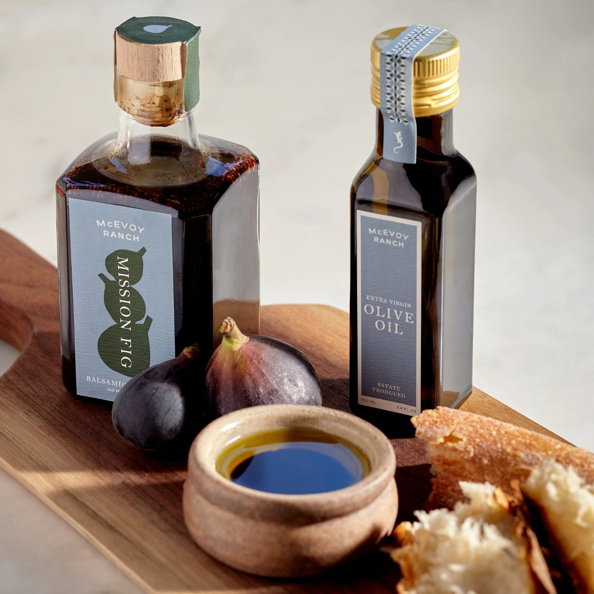 Two bottles of McVoy Ranch olive oil on a wooden board with figs and bread.