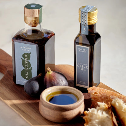 Two bottles of McVoy Ranch olive oil on a wooden board with figs and bread.