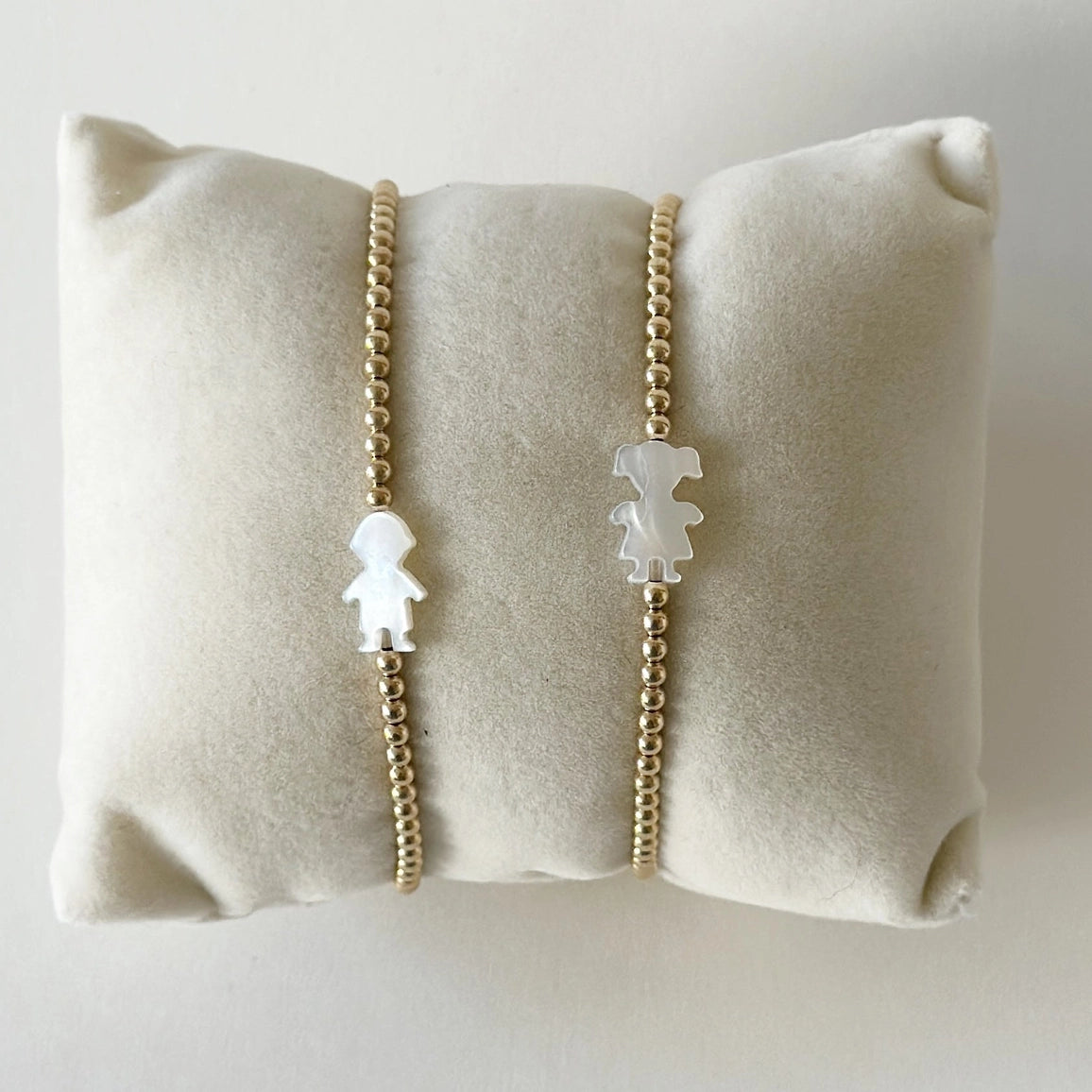Two gold chain bracelets with white character charms on a beige pillow.