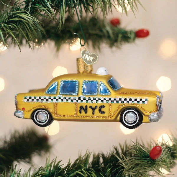 A yellow NYC taxi ornament hanging from a green Christmas tree branch, with blurred holiday lights in the background.