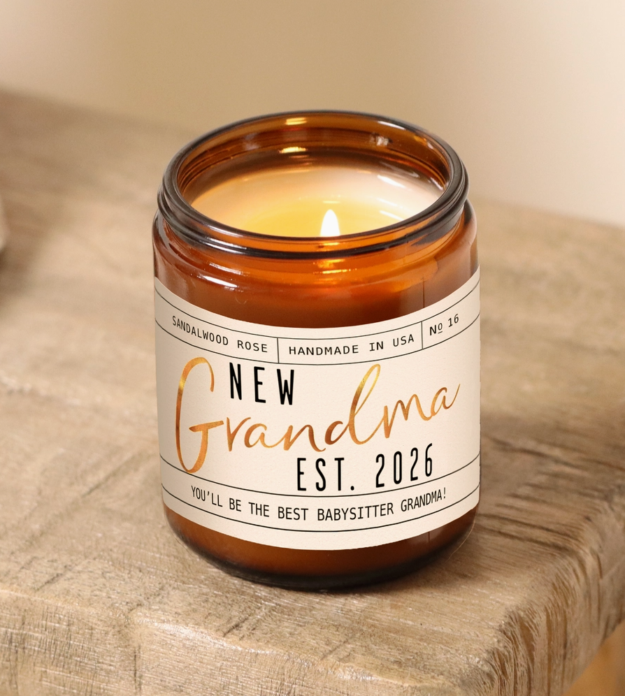 Candle in an amber jar with a label reading 'New Grandma Est. 2026' on a wooden surface.