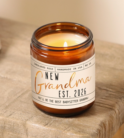 Candle in an amber jar with a label reading 'New Grandma Est. 2026' on a wooden surface.