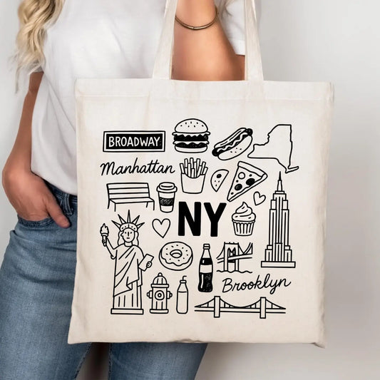A person wearing a white t-shirt and blue jeans holds a cream canvas tote bag printed with black line art icons of NYC landmarks, food, and text including 'Broadway', 'Manhattan', 'NY', and 'Brooklyn'.