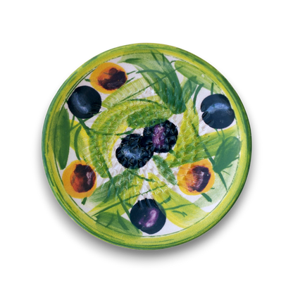 A round ceramic plate featuring a hand-painted design of green leaves, yellow-orange peaches, and dark blue-black plums on a white background, shown from a top-down view against a white backdrop.