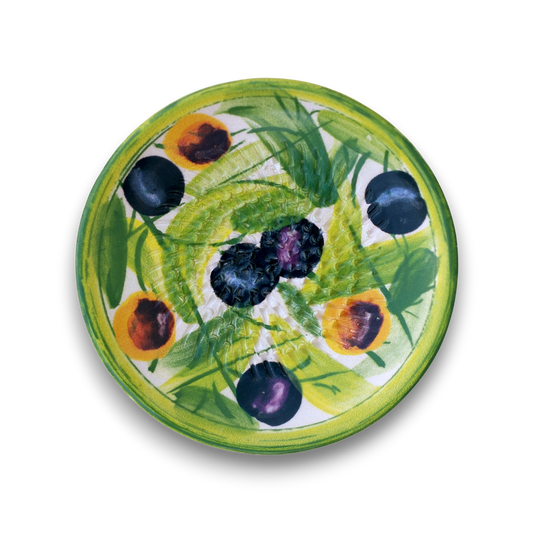 A round ceramic plate featuring a hand-painted design of green leaves, yellow-orange peaches, and dark blue-black plums on a white background, shown from a top-down view against a white backdrop.