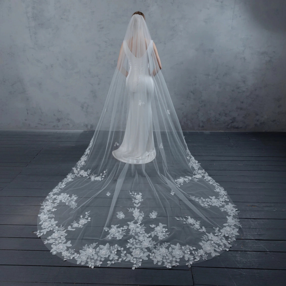 Bride wearing a long, floral lace veil on a dark wooden floor with a gray textured wall background.