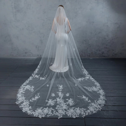 Bride wearing a long, floral lace veil on a dark wooden floor with a gray textured wall background.
