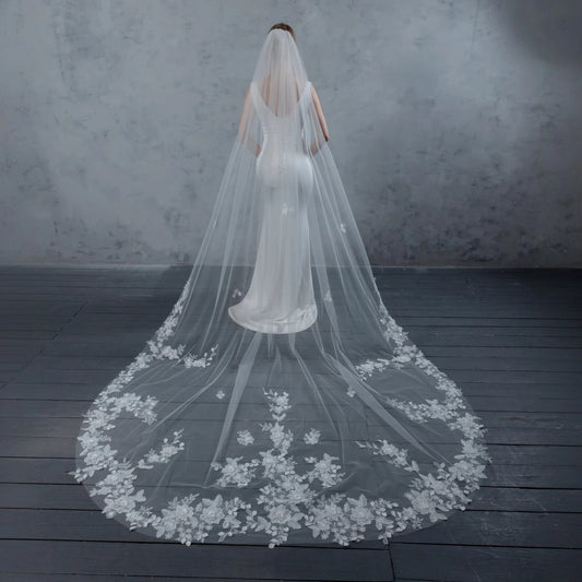 Bride wearing a long, floral lace veil on a dark wooden floor with a gray textured wall background.