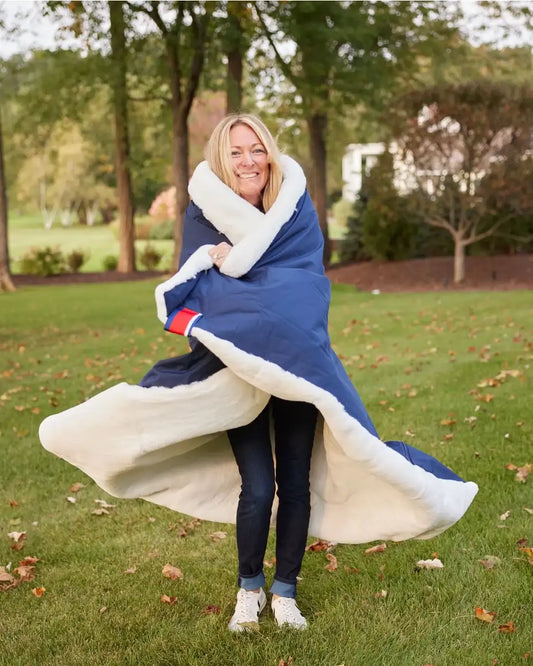 Woman wrapped in a large blue blanket with white trim outdoors on a grassy area.