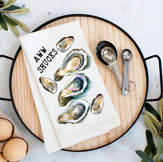 Towel with oyster design and 'Aww Shucks' text on a wooden board with measuring spoons and eggs.