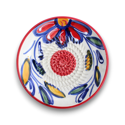 A round white ceramic plate with a hand-painted Talavera style design featuring a red and yellow flower, blue and green leaves, and a textured red spiral in the center, set against a plain white background.