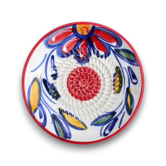 A round white ceramic plate with a hand-painted Talavera style design featuring a red and yellow flower, blue and green leaves, and a textured red spiral in the center, set against a plain white background.