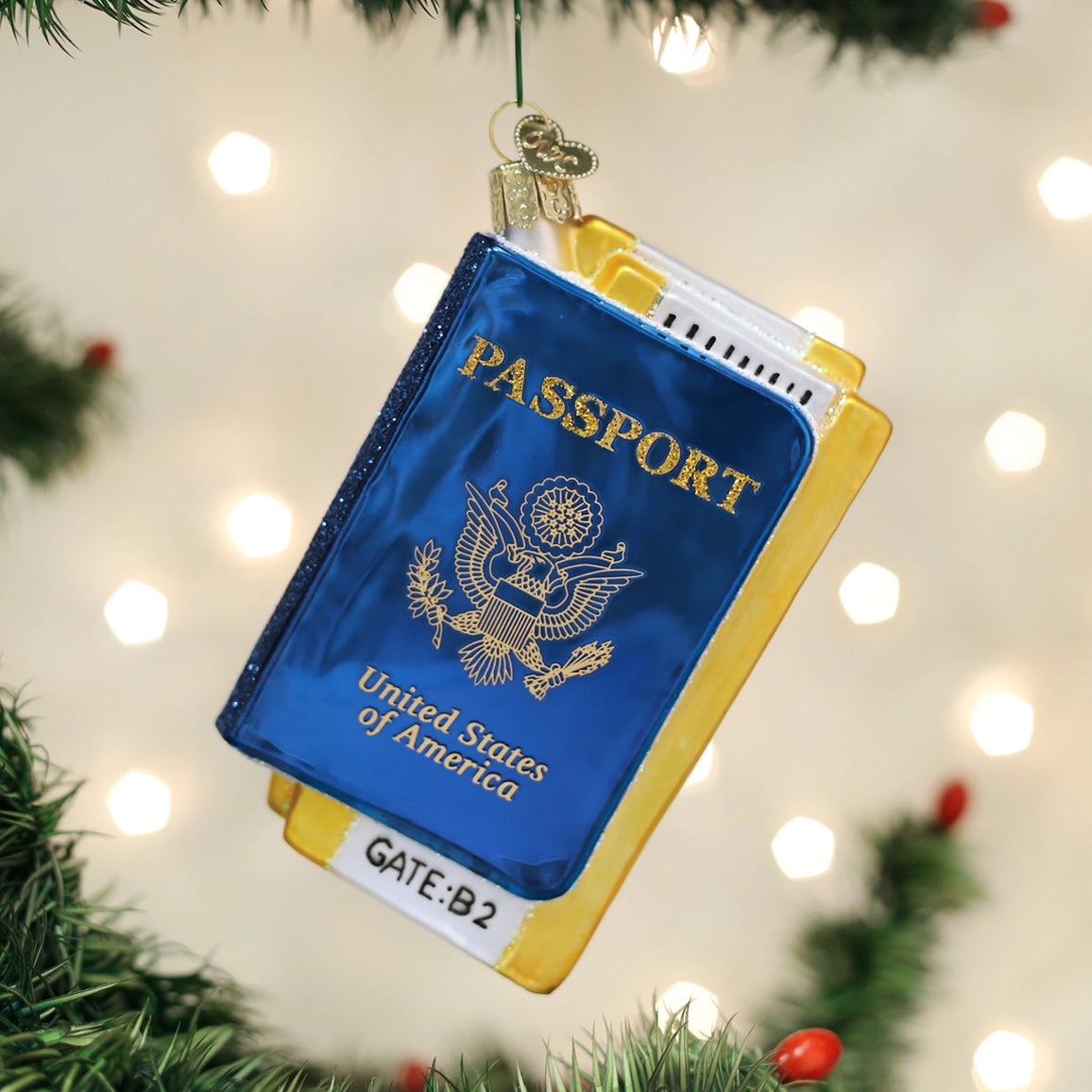 A Christmas ornament shaped like a United States passport hanging from a green Christmas tree branch. The ornament features a blue cover with gold foil-style lettering and emblem, yellow-gold side borders, and a small white label reading 'GATE:B2'.