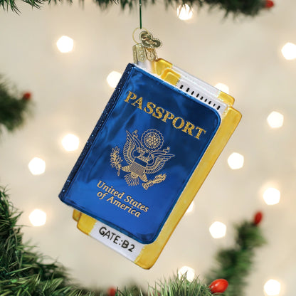 A Christmas ornament shaped like a United States passport hanging from a green Christmas tree branch. The ornament features a blue cover with gold foil-style lettering and emblem, yellow-gold side borders, and a small white label reading 'GATE:B2'.
