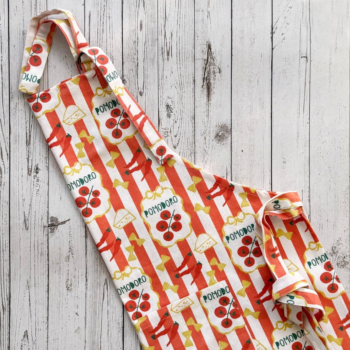 A fabric apron laid flat on a whitewashed wood plank surface. The apron has a repeating pattern of red-and-white vertical stripes, red pom-pom motifs, yellow bows, and the word 'POMODORO" printed in green.