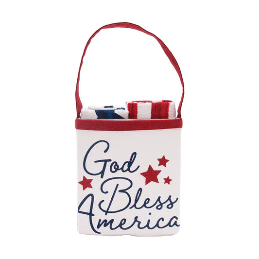 A white fabric tote bag with a red handle, featuring the text 'God Bless America' in navy blue script with red stars on the front. The bag is shown containing folded items, photographed against a solid white background.
