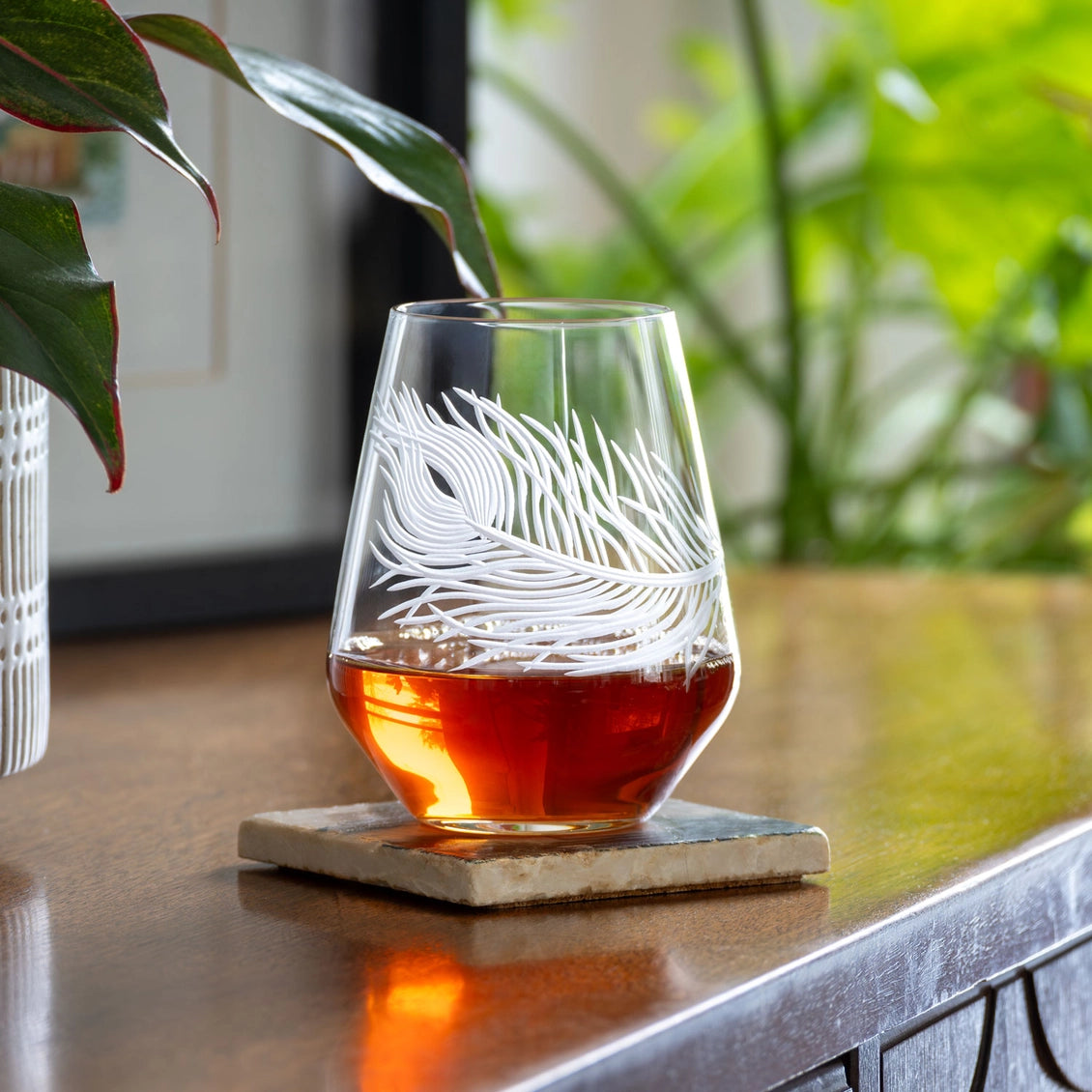 Etched glass with amber liquid on a coaster, surrounded by plants